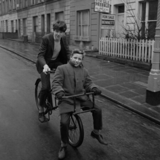 Two teenage boys ride a delivery bike down a terraced residential Lambeth street: one rides the bike and the other sits inside the basket rack, London circa 1964