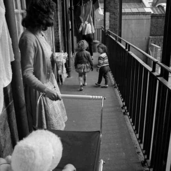 A young woman holds a pram containing a baby wearing a bonnet; she looks away from camera at two small children running along an exterior walkway outside a Peabody block of flats in East London. There is washing drying on lines hanging above the walkway railings in front of the flats; London, circa Spring 1964