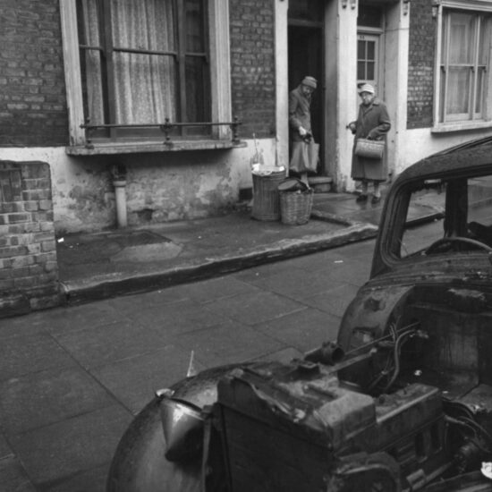Two older women in hats and winter coats stand on the doorstep of a house in a residential street, with a motor car that has been stripped for parts in the foreground, London circa 1964