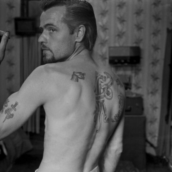 A bearded young man shows off his many traditional tattoos whilst standing bare-chested in Tattoo Bill's parlour, Notting Hill, London circa 1964