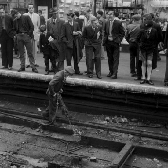 A man in working class clothes operates a jack-hammer on the track at a railway station. He is being observed by men and boys in middle class clothing as they stand on the station platform in front of a tobacconist's shop, circa 1960