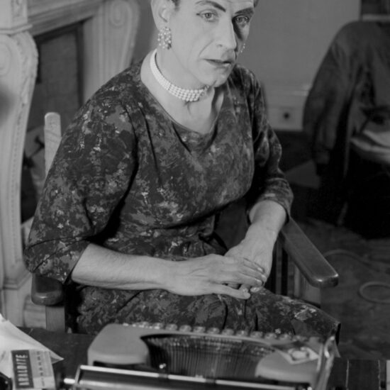 A male transvestite in London's Notting Hill poses in a chair by a typewriter in half drag, including jewellery, dress and make-up, circa 1964