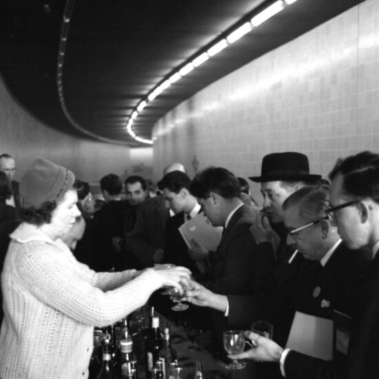 A middle-aged woman in a cardigan and woolly hat stands behind a makeshift bar and serves drinks to men in suits and trilby hats at the opening of the Hyde Park Corner underpass, held on-site, on 17 October 1962 in London. The underpass curves away in the distance and the scene is lit by overhead fluorescent lights
