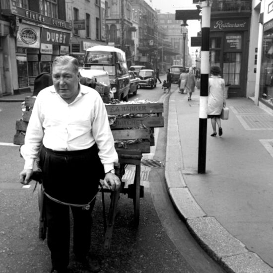 A middle-aged man pulls a handcart into the junction of Wardour Street and Shaftesbury Avenue: the cart carries boxes of vegetables from F A Barker of Datchet. In the background people walk along Wardour Street towards Gerrard Street and vehicles wait at the traffic lights; London, circa Summer 1964
