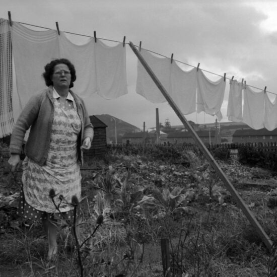 A middle-aged woman wearing a cardigan and an apron over her dress stands by a washing line with Kinglassie colliery in the background. Fife, Scotland, circa 1963